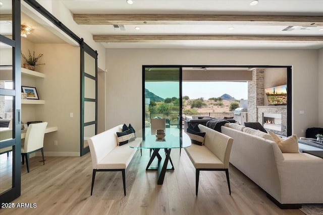 living room with a warm lit fireplace, a barn door, baseboards, light wood-style floors, and beam ceiling