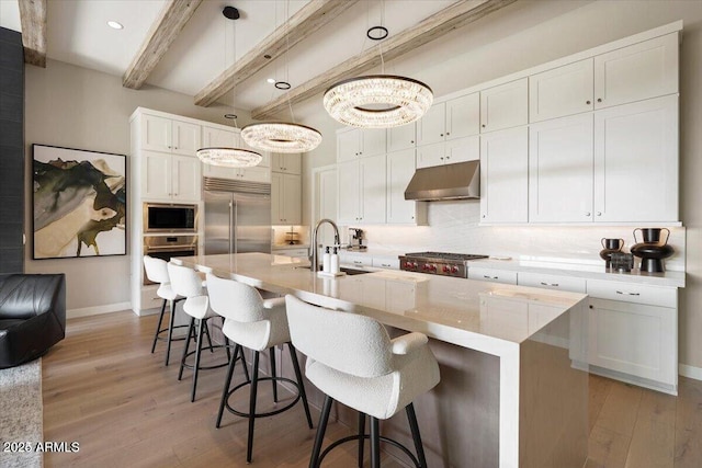 kitchen featuring pendant lighting, light countertops, a kitchen island with sink, built in appliances, and under cabinet range hood