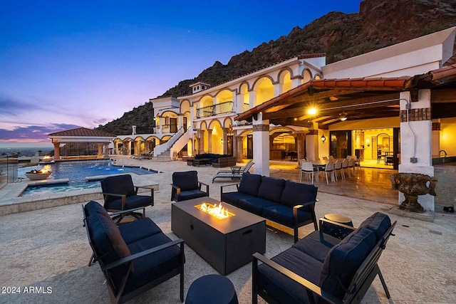 patio terrace at dusk with pool water feature, a mountain view, a swimming pool with hot tub, and an outdoor living space with a fire pit