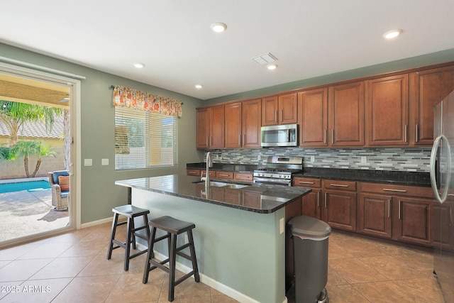 kitchen featuring sink, backsplash, stainless steel appliances, an island with sink, and dark stone counters