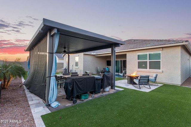 back house at dusk with ceiling fan, an outdoor fire pit, a patio, and a lawn