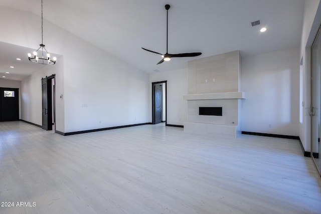 unfurnished living room with ceiling fan with notable chandelier, vaulted ceiling, a barn door, light hardwood / wood-style flooring, and a tiled fireplace