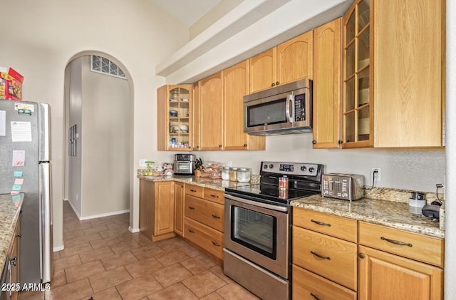 kitchen featuring glass insert cabinets, light stone countertops, appliances with stainless steel finishes, and arched walkways