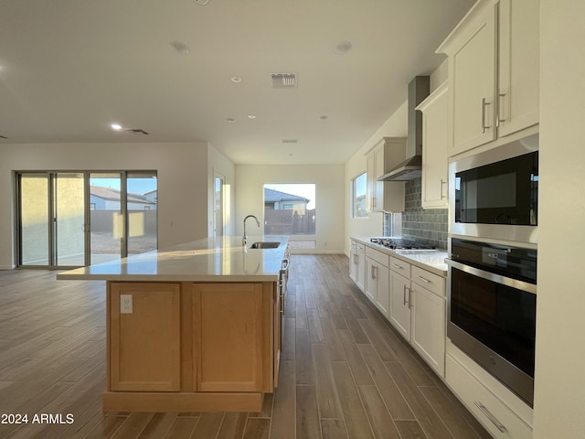 kitchen with appliances with stainless steel finishes, an island with sink, white cabinets, decorative backsplash, and wall chimney exhaust hood