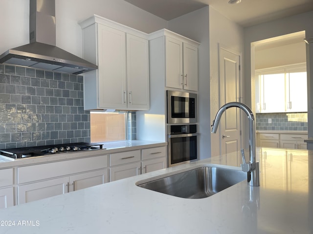 kitchen with sink, white cabinetry, light stone counters, stainless steel appliances, and wall chimney range hood