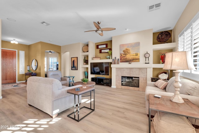 living room featuring ceiling fan, a tiled fireplace, built in features, and light wood-type flooring