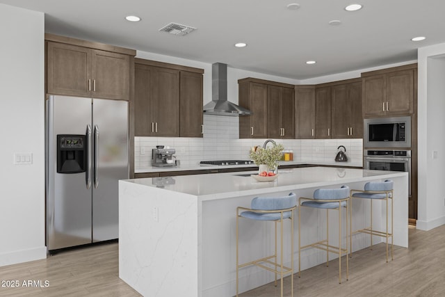 kitchen with wall chimney range hood, appliances with stainless steel finishes, an island with sink, light wood-type flooring, and visible vents