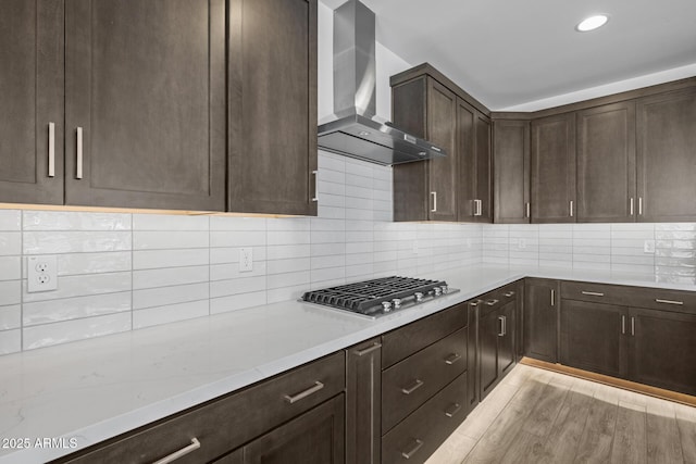 kitchen with wall chimney range hood, dark brown cabinetry, light stone countertops, stainless steel gas cooktop, and decorative backsplash