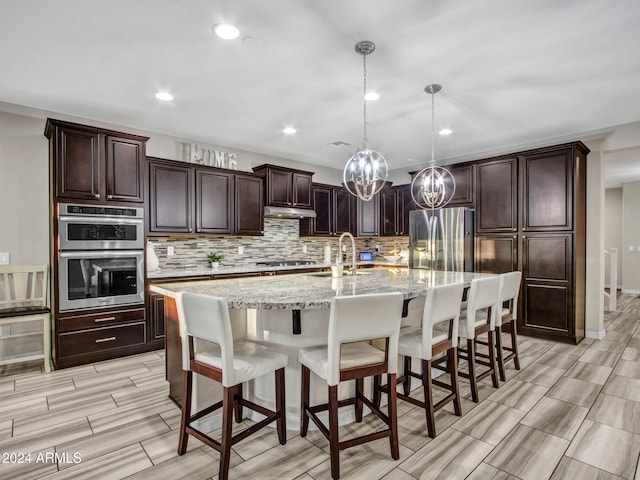 kitchen featuring light stone counters, stainless steel appliances, sink, a center island with sink, and hanging light fixtures