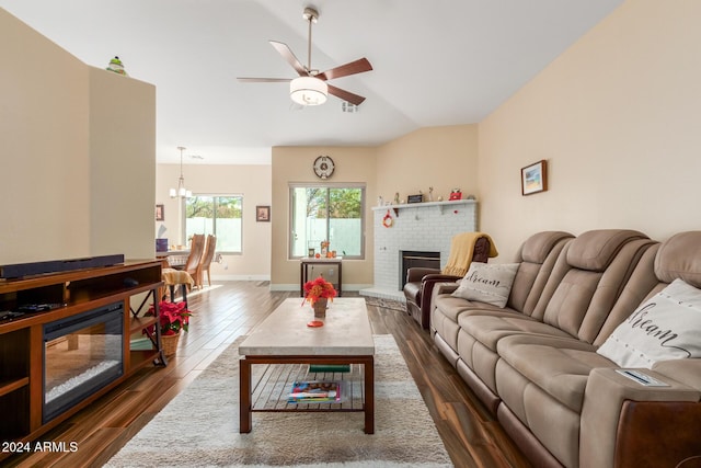 living room with ceiling fan with notable chandelier, lofted ceiling, dark wood-type flooring, and a brick fireplace