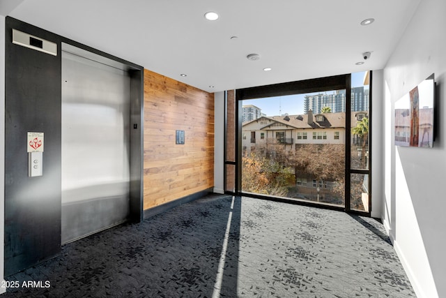 interior space featuring dark colored carpet, elevator, a wall of windows, and wood walls