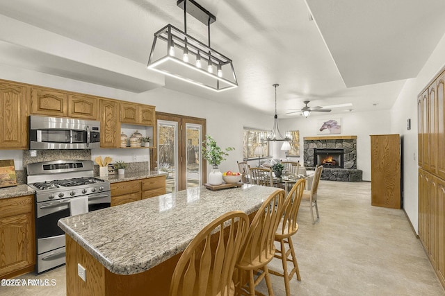 kitchen with a fireplace, appliances with stainless steel finishes, a center island, brown cabinets, and ceiling fan with notable chandelier