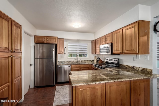 kitchen with stainless steel appliances, a peninsula, a sink, brown cabinetry, and dark stone countertops