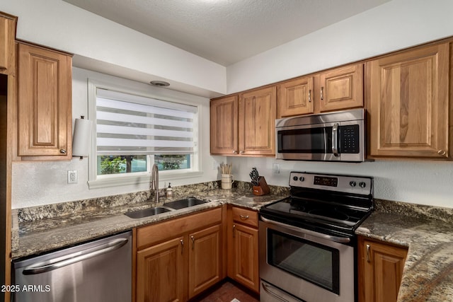 kitchen featuring brown cabinetry, dark stone counters, stainless steel appliances, and a sink