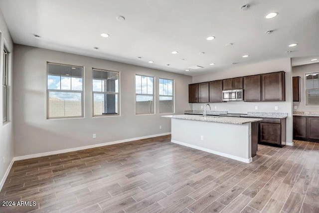 kitchen with light stone counters, dark brown cabinetry, sink, wood-type flooring, and an island with sink