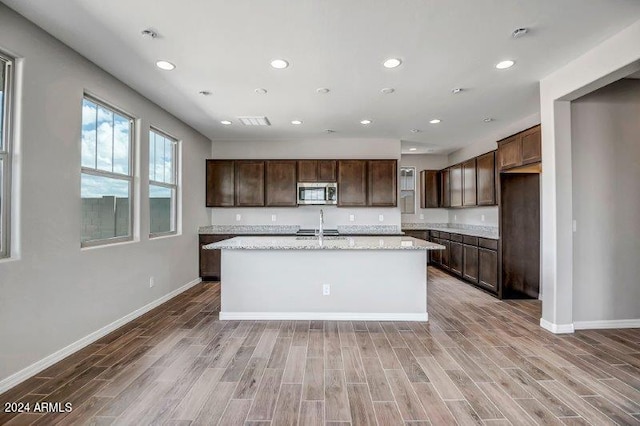 kitchen with hardwood / wood-style floors, dark brown cabinets, light stone counters, and a kitchen island with sink