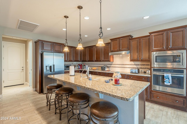 kitchen with backsplash, an island with sink, decorative light fixtures, light stone counters, and stainless steel appliances