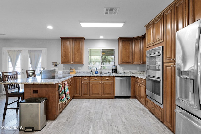 kitchen with a kitchen breakfast bar, brown cabinetry, appliances with stainless steel finishes, light stone countertops, and a peninsula
