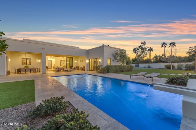 pool at dusk featuring ceiling fan, fence, a lawn, outdoor lounge area, and a patio area