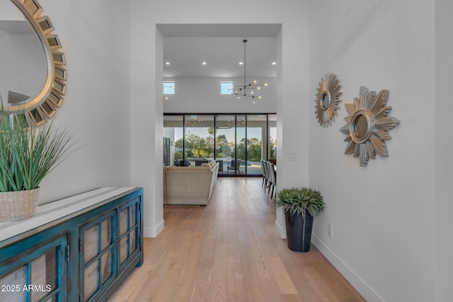 hallway with light wood-style flooring, a notable chandelier, baseboards, and a towering ceiling