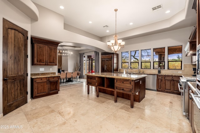 kitchen featuring an island with sink, hanging light fixtures, a notable chandelier, light stone counters, and stainless steel appliances