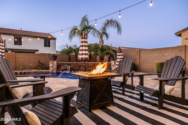 patio terrace at dusk with a fenced in pool, pool water feature, and an outdoor fire pit