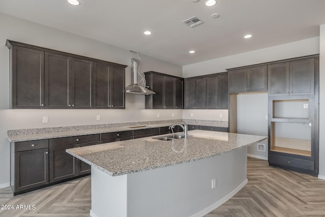 kitchen featuring an island with sink, light stone counters, wall chimney exhaust hood, light parquet flooring, and sink