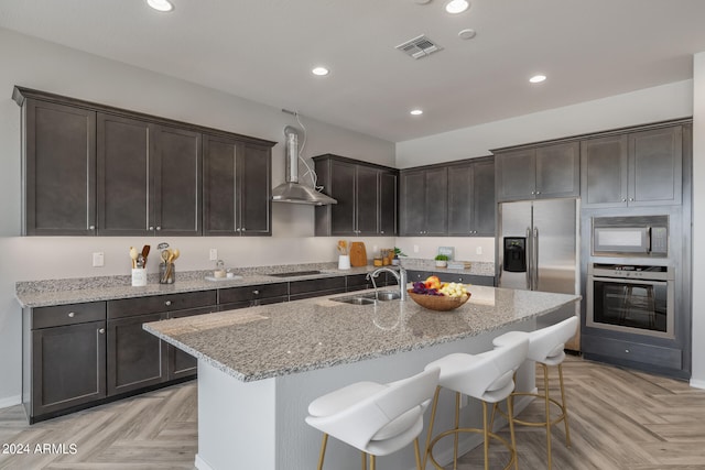 kitchen with light parquet floors, a kitchen island with sink, black appliances, wall chimney range hood, and a breakfast bar area