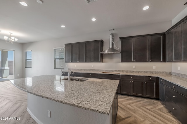 kitchen featuring wall chimney range hood, a kitchen island with sink, light stone counters, light parquet flooring, and sink