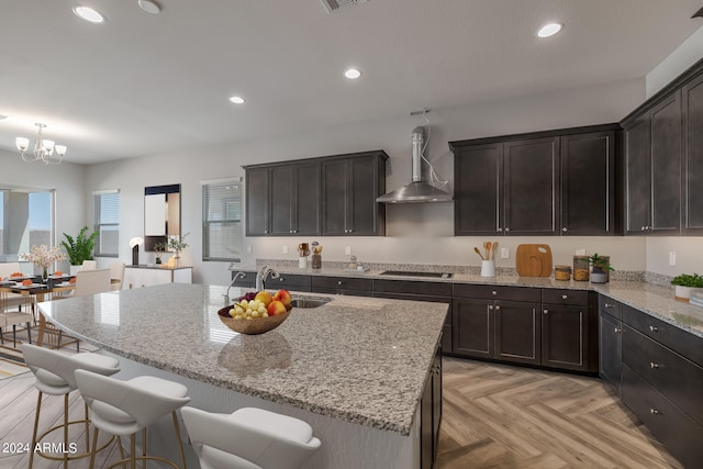 kitchen with light stone counters, light parquet floors, an island with sink, wall chimney exhaust hood, and sink