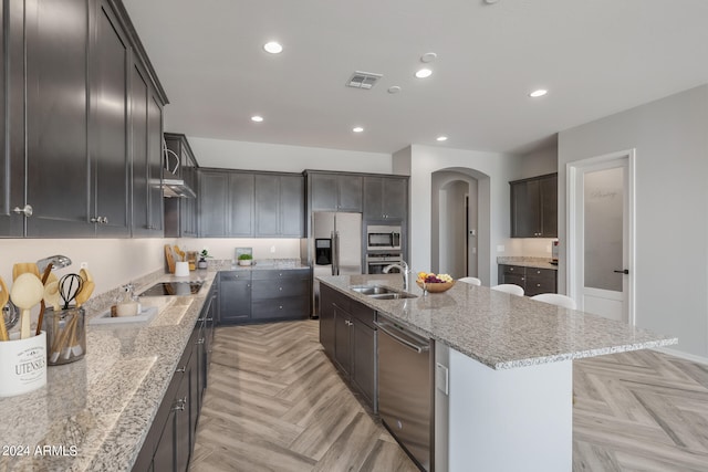 kitchen with an island with sink, appliances with stainless steel finishes, light stone counters, light parquet floors, and sink