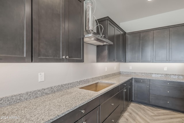 kitchen with wall chimney range hood, light stone countertops, dark brown cabinets, and light parquet flooring