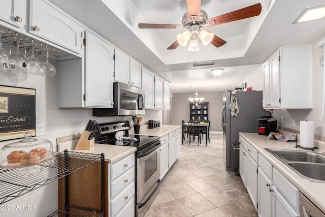 kitchen with ceiling fan with notable chandelier, a raised ceiling, sink, white cabinetry, and stainless steel appliances