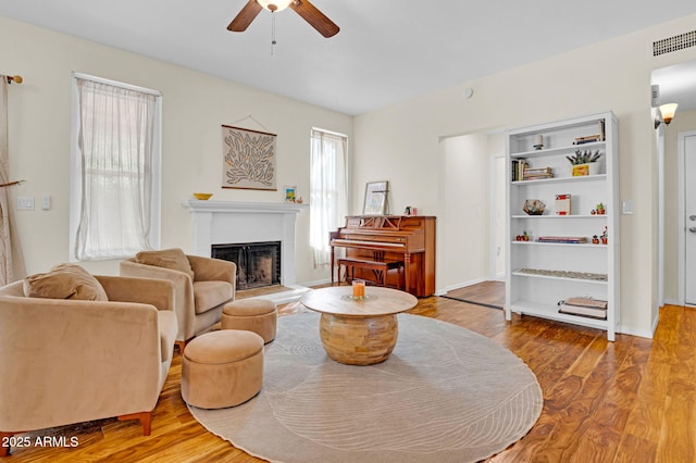 living area featuring a ceiling fan, wood finished floors, a fireplace with raised hearth, baseboards, and visible vents