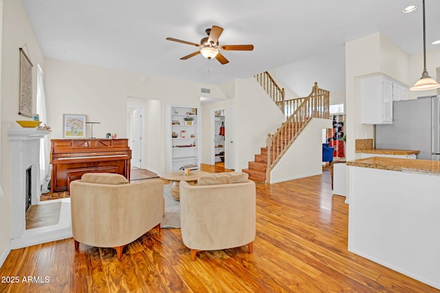 living room featuring light wood-type flooring, a ceiling fan, a fireplace with raised hearth, and stairs