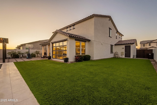 back of house at dusk with stucco siding, a lawn, a patio area, fence, and a tile roof