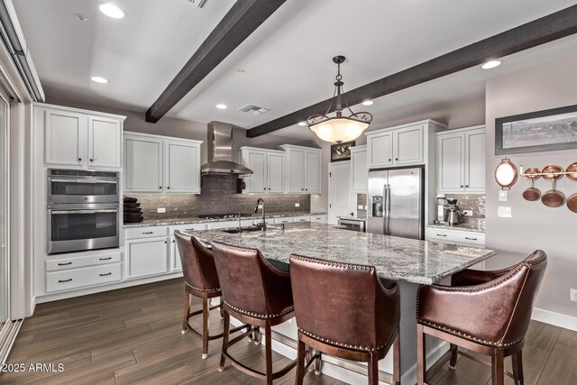 kitchen featuring a kitchen island with sink, wall chimney range hood, appliances with stainless steel finishes, and white cabinetry