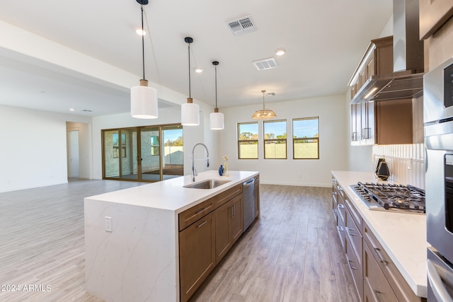 kitchen featuring pendant lighting, wall chimney range hood, stainless steel appliances, sink, and a center island with sink