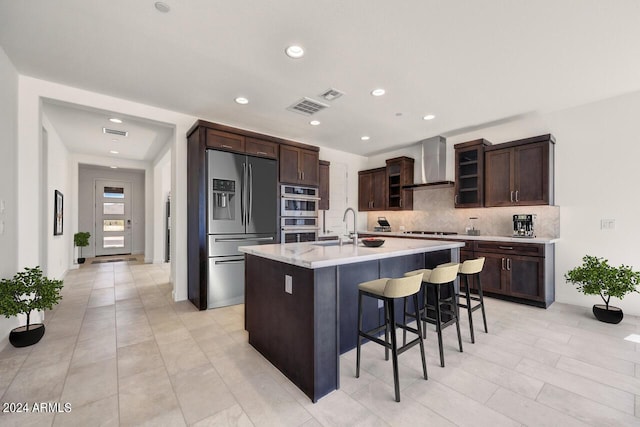 kitchen featuring sink, wall chimney range hood, a kitchen island with sink, dark brown cabinets, and appliances with stainless steel finishes