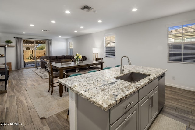 kitchen with sink, gray cabinetry, stainless steel dishwasher, light stone countertops, and a center island with sink