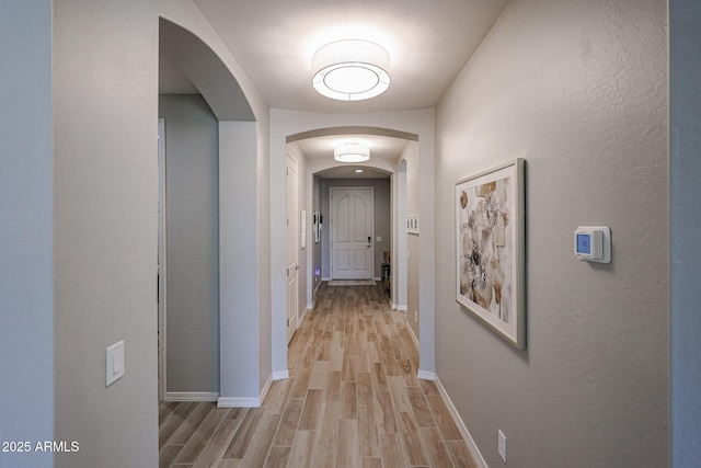 corridor featuring arched walkways, light wood finished floors, and baseboards