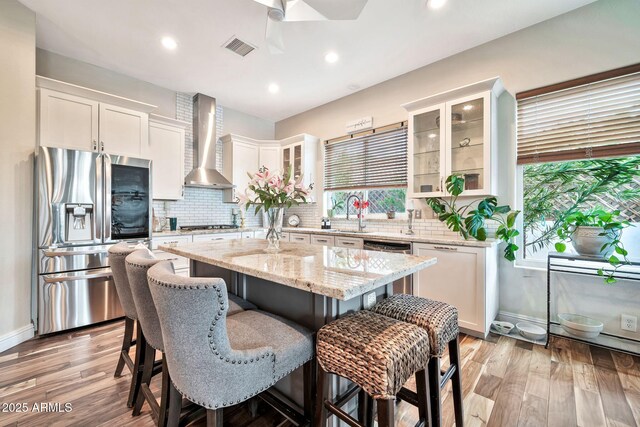 kitchen featuring appliances with stainless steel finishes, white cabinetry, a center island, light stone counters, and wall chimney exhaust hood