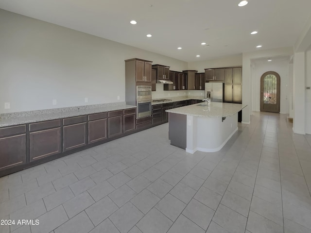 kitchen with a center island with sink, sink, light stone countertops, light tile patterned floors, and appliances with stainless steel finishes