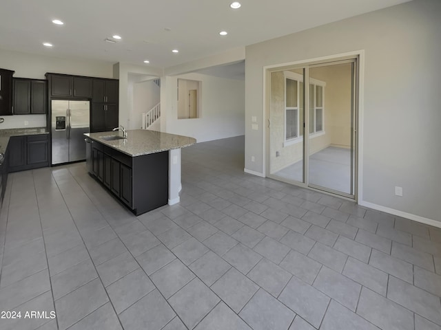 kitchen with sink, light stone counters, stainless steel fridge, a kitchen island with sink, and light tile patterned flooring