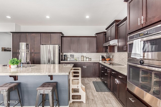 kitchen with a breakfast bar area, light stone counters, dark brown cabinets, stainless steel appliances, and backsplash