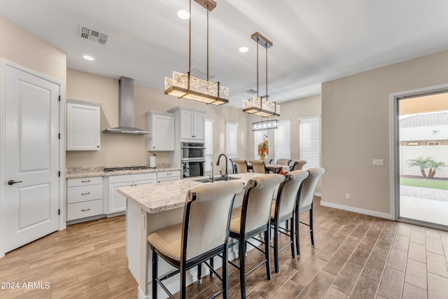 kitchen with a kitchen breakfast bar, wall chimney exhaust hood, pendant lighting, a center island with sink, and white cabinetry