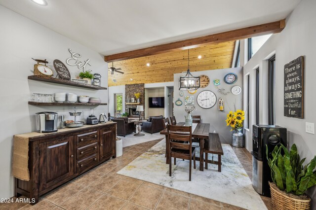 tiled dining space featuring beamed ceiling, ceiling fan with notable chandelier, and a stone fireplace