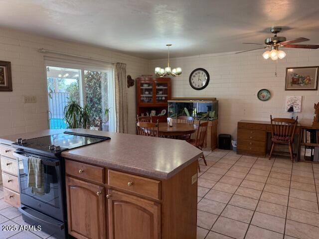 kitchen with brown cabinetry, electric range, light tile patterned flooring, ceiling fan with notable chandelier, and a center island