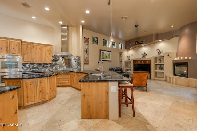 kitchen featuring ceiling fan, wall chimney exhaust hood, dark stone counters, a kitchen island with sink, and a fireplace