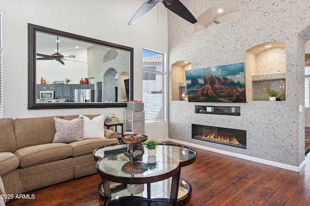 living room with wood-type flooring, a high ceiling, a glass covered fireplace, ceiling fan, and baseboards